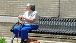 Older lady waiting on a bench alone and with a cane.